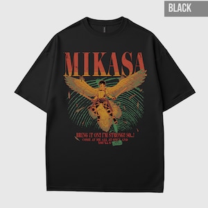 Puede incluir: Camiseta negra con la palabra "MIKASA" en naranja y amarillo, y un gráfico de un personaje alado. El texto "BRING IT ON!! I'M STRONG!! SO...! COME AT ME ALL AT ONCE AND YOU'LL S..." está impreso en la camiseta.