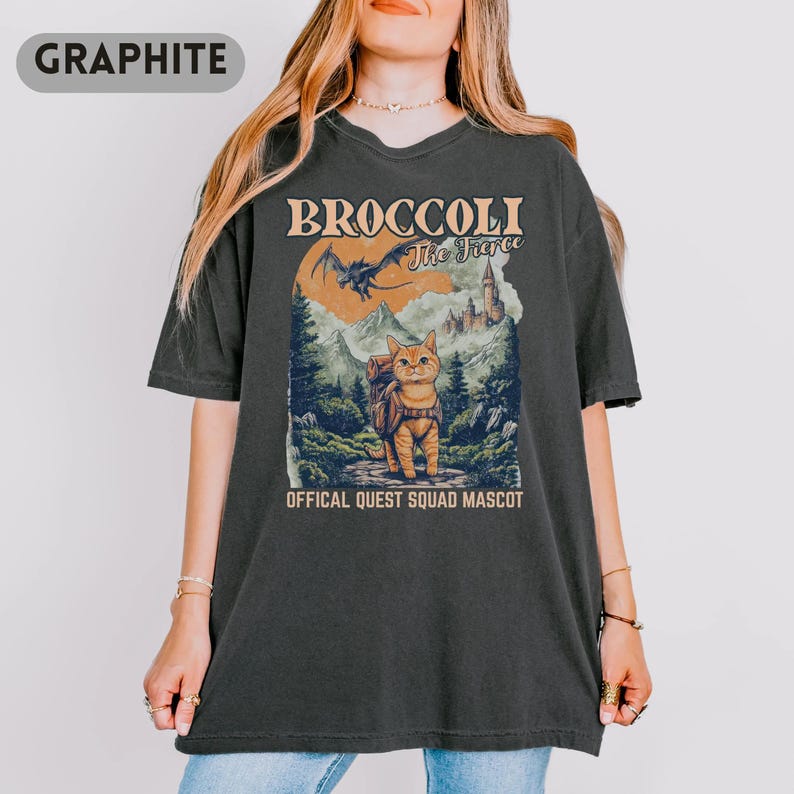 May include: Graphite-colored t-shirt featuring a graphic of a cat with a backpack in a fantasy landscape. The text reads "BROCCOLI The Fierce" above the image, with "OFFICAL QUEST SQUAD MASCOT" below. The design includes a dragon, mountains, and a castle.