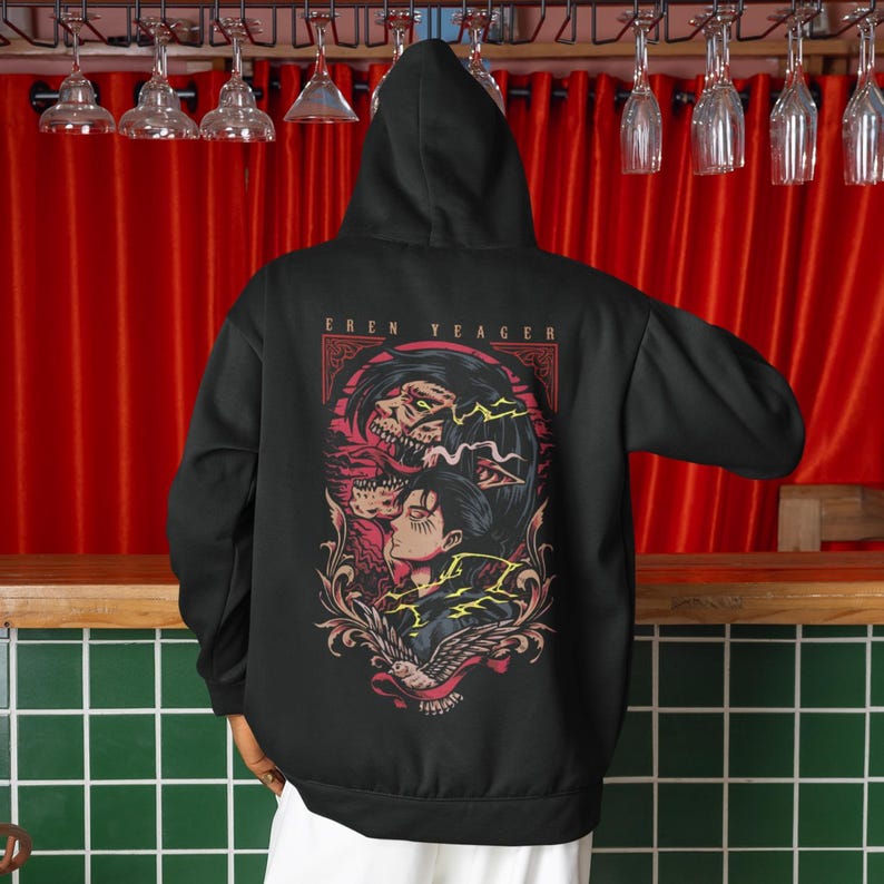 Puede incluir: Una sudadera con capucha negra que presenta un estampado gr&aacute;fico en la parte posterior. El dise&ntilde;o muestra una ilustraci&oacute;n estilizada de Eren Yeager con una forma de tit&aacute;n arriba, rayos amarillos y un p&aacute;jaro abajo, con el texto EREN YEAGER impreso.