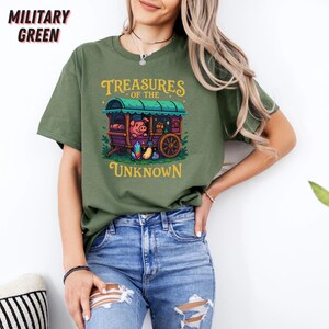 May include: Military green t-shirt with a colorful graphic of a pig in a wagon, with the text "Treasures of the Unknown" in gold lettering. The shirt is made of a soft material and has short sleeves.
