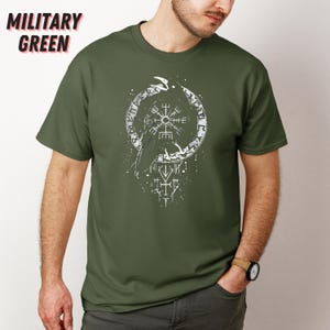 K&ouml;nnte beinhalten: Milit&auml;rgr&uuml;nes T-Shirt mit einem wei&szlig;en Grafikdesign eines Raben in einem kreisf&ouml;rmigen Rahmen mit Runen. Der Text "MILITARY GREEN" befindet sich oben links. Das Shirt besteht aus einem weichen, bequemen Material.