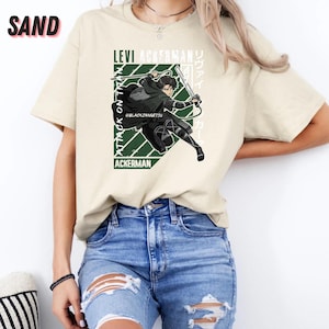 May include: Sand-colored t-shirt featuring a graphic of a character from "Attack on Titan" with the text "Levi Ackerman" and Japanese text. The character is depicted in action with a sword, set against a green and white geometric design.