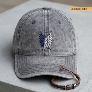 May include: A charcoal grey baseball cap with a washed, distressed finish. The front features an embroidered emblem of two stylised wings, one in blue and one in white. A brown leather cord bracelet with metallic beads rests near the brim.