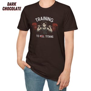 K&ouml;nnte beinhalten: Dunkelbraunes T-Shirt mit dem Text "TRAINING TO KILL TITANS" und einer Grafik eines Gewichthebers. Die Langhantel hat rote Gewichte. Die Worte "DARK CHOCOLATE" sind oben links aufgedruckt.
