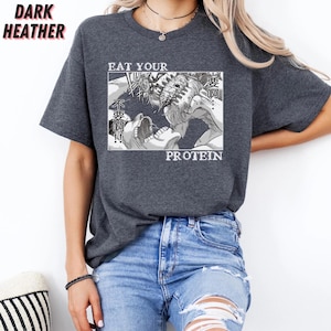 May include: Dark heather gray t-shirt with a manga-style graphic. The graphic features a black and white illustration of a battle scene with the words "EAT YOUR PROTEIN" in white text. The shirt has short sleeves and a crew neck.
