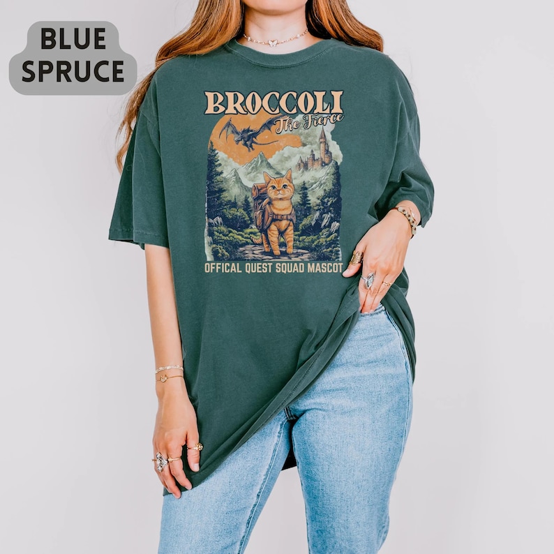 May include: Blue Spruce oversized t-shirt featuring a graphic of a cat with a backpack, mountains, and a dragon. The text on the shirt reads "BROCCOLI The Fierce" and "OFFICAL QUEST SQUAD MASCOT".
