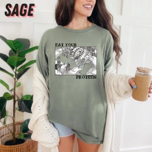May include: Sage green t-shirt with a black and white manga-style graphic. The graphic features two figures in a battle scene, with the words "EAT YOUR PROTEIN" and Japanese text. The word "SAGE" is printed in pink at the top.