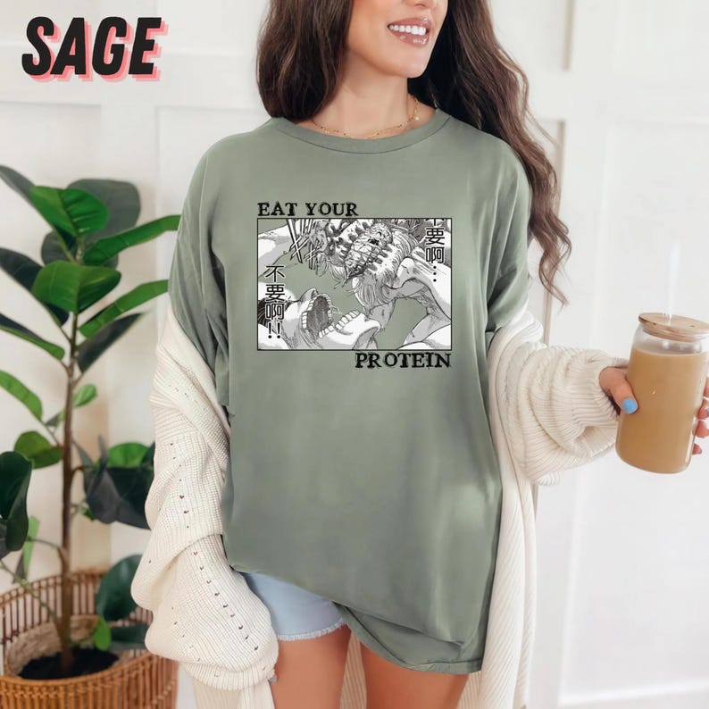 May include: Sage green t-shirt with a black and white manga-style graphic. The graphic features two figures in a battle scene, with the words "EAT YOUR PROTEIN" and Japanese text. The word "SAGE" is printed in pink at the top.