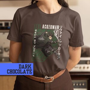 May include: Dark chocolate t-shirt featuring a graphic of Levi Ackerman from Attack on Titan. The design includes text in English and Japanese, with a green and white color scheme. The shirt is a crew neck.