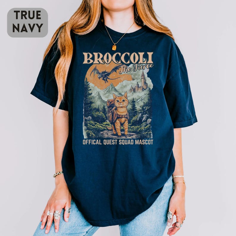 May include: True navy blue t-shirt with a graphic featuring a cat with a backpack in a forest scene, the text "BROCCOLI The Fierce" and "OFFICAL QUEST SQUAD MASCOT". The shirt is a comfortable, casual style.