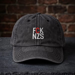 Puede incluir: Una gorra de béisbol gris carbón con un acabado desgastado y lavado. La parte frontal presenta un texto bordado en blanco que dice FCK NZS, con la letra C cosida en un hilo rojo contrastante. La gorra tiene una visera curva y seis paneles.