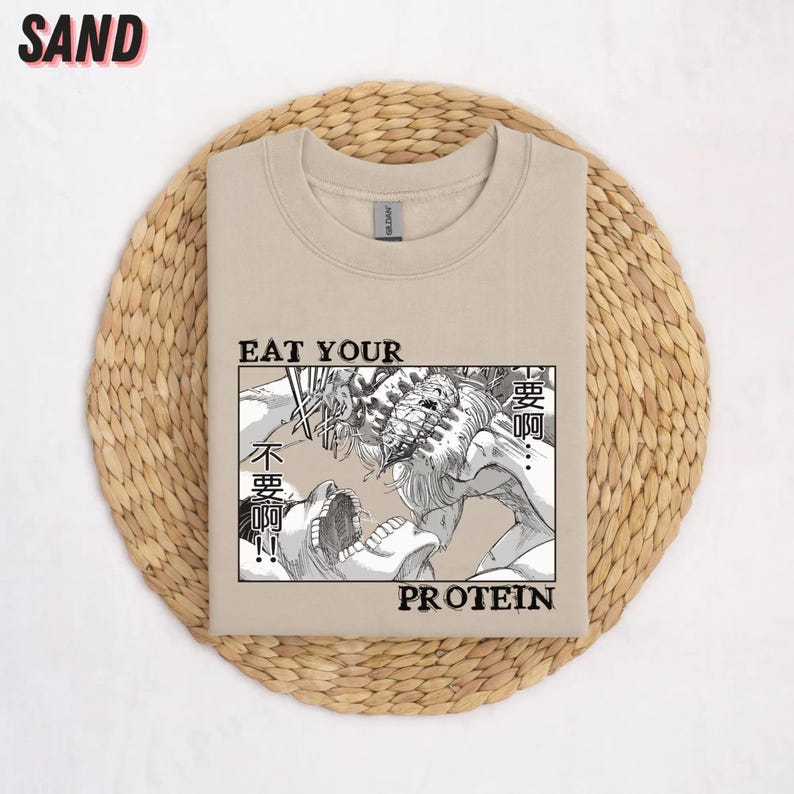 May include: Sand-colored sweatshirt with a graphic featuring a manga-style illustration of two figures in a battle scene. The text "EAT YOUR PROTEIN" is displayed below the illustration, with Japanese text above.