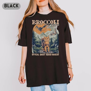 May include: Black t-shirt featuring a graphic of a cat with a backpack, mountains, and a castle. Text on the shirt reads "BROCCOLI The Fierce" and "OFFICAL QUEST SQUAD MASCOT". The design has a vintage, outdoorsy aesthetic.