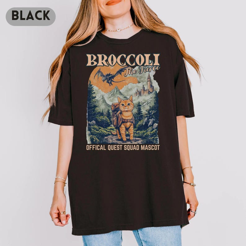 May include: Black t-shirt featuring a graphic of a cat with a backpack, mountains, and a castle. Text on the shirt reads "BROCCOLI The Fierce" and "OFFICAL QUEST SQUAD MASCOT". The design has a vintage, outdoorsy aesthetic.