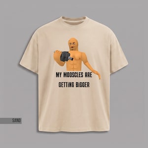 Op de afbeelding: Een zandkleurig t-shirt met een afbeelding van een gespierd, lichtgetint personage dat een donkergrijs object vasthoudt. Onder het personage staat de zwarte tekst: MY MOOSCLES ARE GETTING BIGGER. Het t-shirt heeft korte mouwen en een ronde hals.
