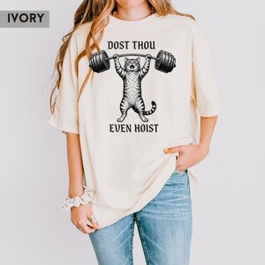 May include: Ivory-coloured t-shirt with a black and white illustration of a cat lifting a barbell. The text "DOST THOU EVEN HOIST" is printed above and below the cat graphic. The shirt has a relaxed fit.
