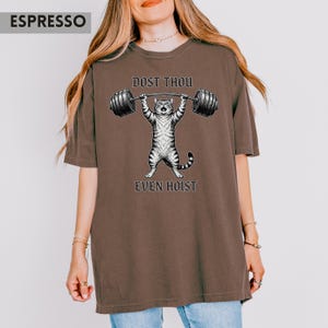 May include: Espresso-coloured t-shirt with a cartoon cat lifting a barbell. The cat is grey and white, with the words "DOST THOU" above the barbell and "EVEN HOIST" below. The shirt has a relaxed fit.