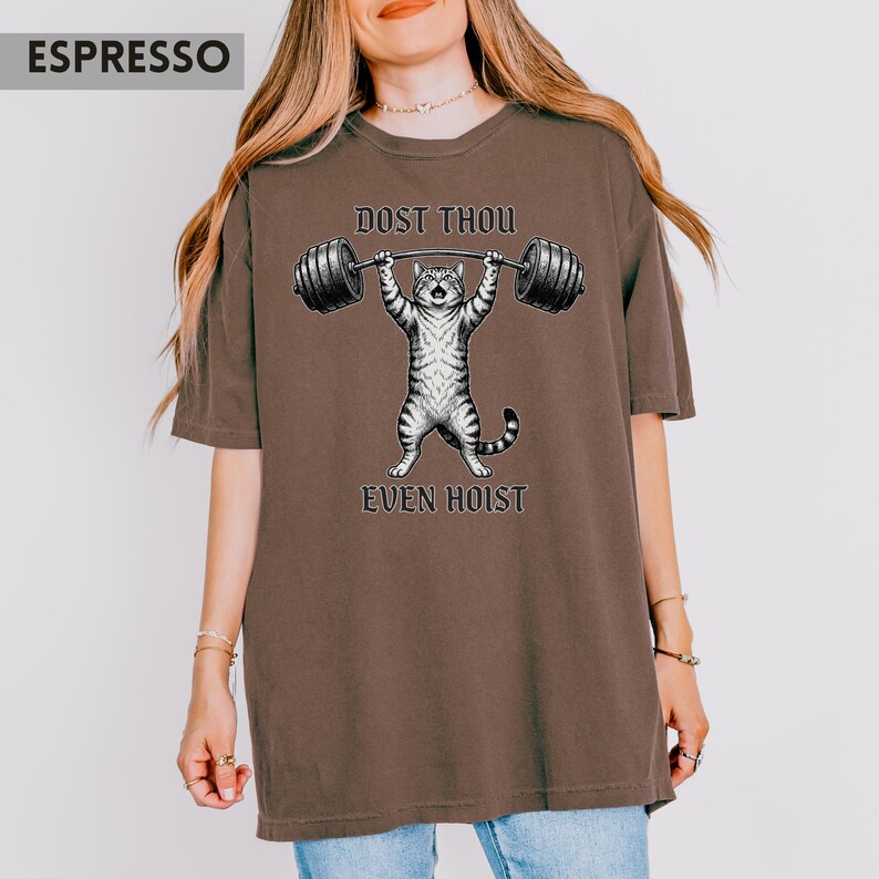 May include: Espresso-coloured t-shirt with a cartoon cat lifting a barbell. The cat is grey and white, with the words "DOST THOU" above the barbell and "EVEN HOIST" below. The shirt has a relaxed fit.