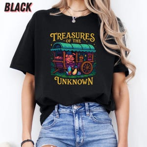 May include: Black t-shirt featuring a whimsical illustration of a pig in a caravan with the text "Treasures of the Unknown". The design includes a green roof, purple wagon, and colorful gems. The word "BLACK" is printed at the top.
