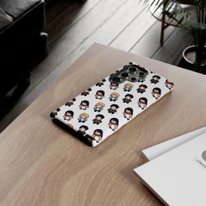 Peut inclure: Une coque de t&eacute;l&eacute;phone blanche avec un motif r&eacute;p&eacute;t&eacute; de personnages de dessins anim&eacute;s en diff&eacute;rentes tenues. L'&eacute;tui a une d&eacute;coupe pour l'appareil photo et repose sur une surface en bois. Le motif comprend des personnages avec diff&eacute;rentes couleurs de cheveux et v&ecirc;tements.