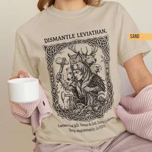 May include: Sand-coloured t-shirt with a detailed black graphic. The design includes a central figure and animals, with the text "DISMANTLE LEVIATHAN." Below, it reads "Learn survival skills. Honour the Earth. Destroy Civilization. Bring about sustainability. Go FERAL."