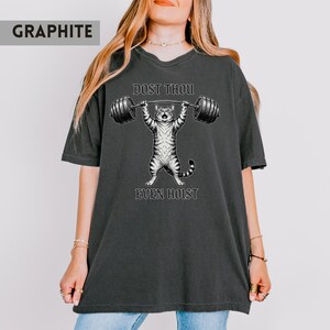 May include: Graphite-coloured t-shirt with a cartoon cat lifting weights. The black and white cat illustration is between the text "DOST THOU" and "EVEN HOIST". The shirt has short sleeves and is a dark grey colour.