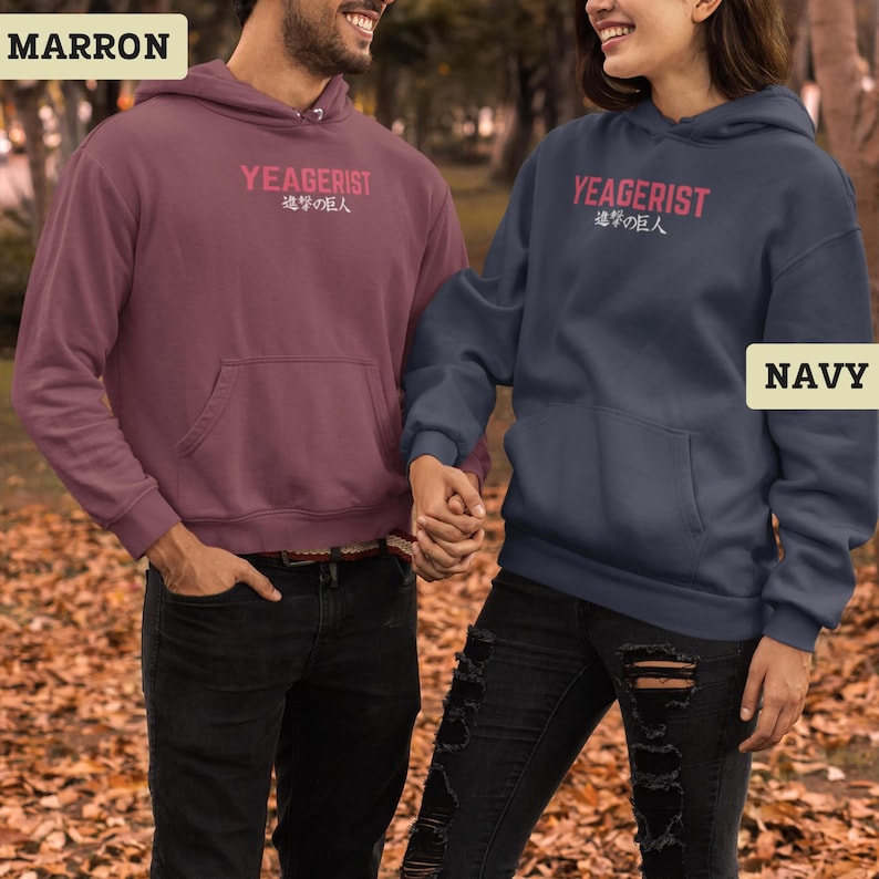 Puede incluir: Una sudadera con capucha color granate y una sudadera con capucha azul marino, ambas con bolsillo tipo canguro, mangas largas y capucha. Cada prenda muestra el texto YEAGERIST en letras rosas sobre caracteres japoneses blancos en el pecho.