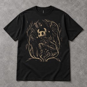 May include: A black short-sleeved t-shirt featuring a tan-coloured graphic print of a horned character standing on a rock formation, surrounded by stylised vines and falling leaves. The design is centred on the chest of the garment.