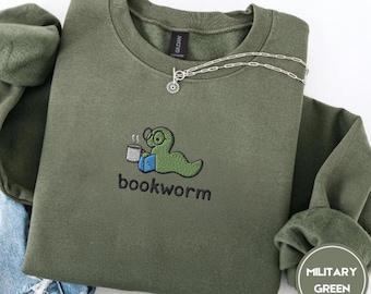 May include: Olive green sweatshirt with an embroidered "bookworm" design. The design features a green worm wearing glasses, holding a mug and a book. A silver necklace is draped across the sweatshirt. The words "Military Green" are in a circle in the bottom right corner.