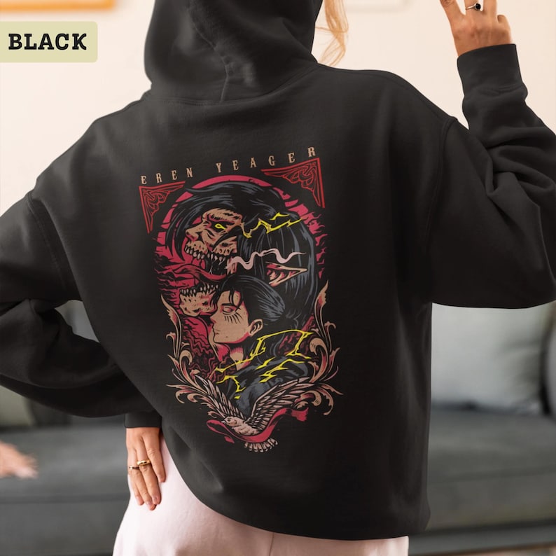Puede incluir: Una sudadera con capucha negra que presenta un gr&aacute;fico en la parte posterior con el texto EREN YEAGER. El dise&ntilde;o muestra una ilustraci&oacute;n estilizada de un personaje y una figura monstruosa con acentos rojos y amarillos, enmarcada por volutas.