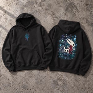 Op de afbeelding: Een zwarte hoodie met een kleine blauwe en rode afbeelding op de borst. De achterkant toont een grote, gedetailleerde illustratie van twee figuren met witte maskers, omringd door blauwe vlinders en donker gebladerte in een bos.