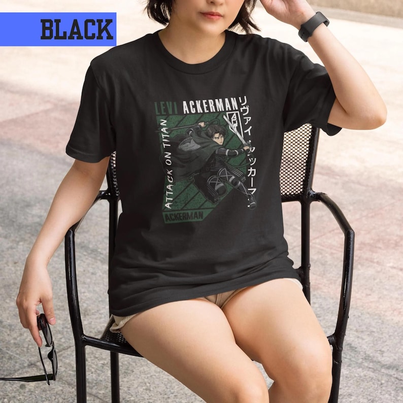 May include: Black t-shirt featuring a graphic of a character from "Attack on Titan" with the text "Levi Ackerman" and Japanese text. The design includes a green geometric pattern and the words "Attack on Titan" in white. The word "BLACK" is in a blue rectangle.