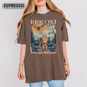 May include: Espresso-colored t-shirt featuring a graphic of a cat with a backpack in a forest scene. The text reads "BROCCOLI The Fierce" and "OFFICAL QUEST SQUAD MASCOT". The design includes a dragon, mountains, and a castle.
