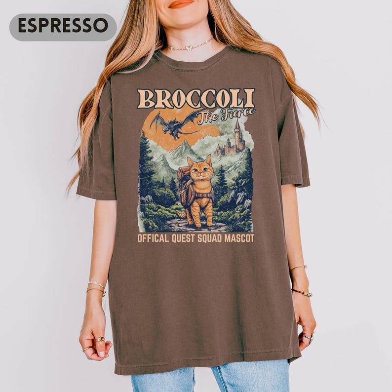 May include: Espresso-colored t-shirt featuring a graphic of a cat with a backpack in a forest scene. The text reads "BROCCOLI The Fierce" and "OFFICAL QUEST SQUAD MASCOT". The design includes a dragon, mountains, and a castle.