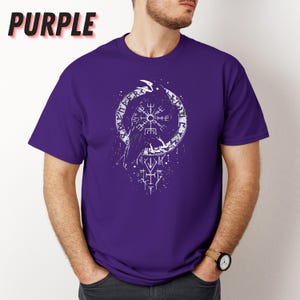 K&ouml;nnte beinhalten: Ein lila T-Shirt mit einer wei&szlig;en Grafik eines Raben und Runen in einem Kreis. Das Wort "PURPLE" ist oben links in Schwarz gedruckt. Das T-Shirt hat kurze &Auml;rmel und einen Rundhalsausschnitt. Eine Uhr wird getragen.