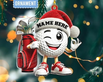 Puede incluir: Un adorno plano 2D con un personaje de pelota de golf guiñando un ojo y usando un gorro de Papá Noel con "NAME HERE" impreso. La pelota de golf sostiene una bolsa de golf roja con palos y usa zapatillas rojas y blancas. El adorno cuelga de un hilo dorado.