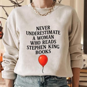 May include: A beige sweatshirt with the text "NEVER UNDERESTIMATE A WOMAN WHO READS STEPHEN KING BOOKS" in black. A red balloon graphic is below the text. The sweatshirt is a casual style.