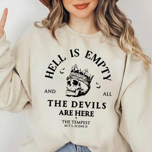 Op de afbeelding: Crèmekleurige sweatshirt met de zwarte tekst "HELL IS EMPTY AND ALL THE DEVILS ARE HERE", een gekroonde schedel en de tekst "THE TEMPEST ACT I, SCENE II".