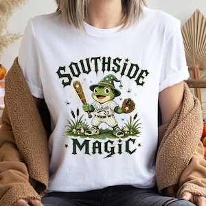 May include: White t-shirt featuring a cartoon frog wearing a baseball uniform and a wizard hat, holding a bat and glove. The shirt has the words "Southside Magic" in green lettering. The frog is standing in a field of grass and mushrooms.