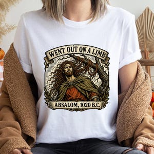 Può includere: T-shirt bianco con un'illustrazione dettagliata di un uomo con capelli lunghi e barba, che indossa un mantello. La grafica include il testo "WENT OUT ON A LIMB" e "ABSALOM, 1020 B.C." in stile vintage.
