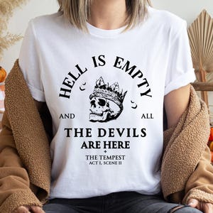 May include: White t-shirt with a black graphic of a crowned skull and the text "HELL IS EMPTY AND ALL THE DEVILS ARE HERE" from Shakespeare's "The Tempest". The shirt also includes the text "ACT I, SCENE II".