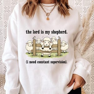 May include: White sweatshirt with the text "the lord is my shepherd. (i need constant supervision)." The graphic shows cartoon sheep behind a wooden fence with birds perched on top.