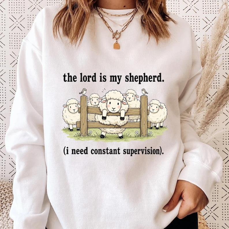 May include: White sweatshirt with the text "the lord is my shepherd. (i need constant supervision)." The graphic shows cartoon sheep behind a wooden fence with birds perched on top.