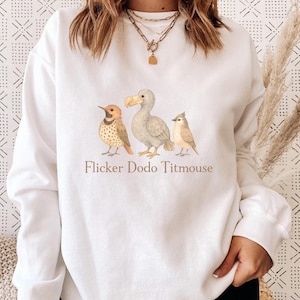 May include: White sweatshirt featuring a watercolor illustration of three birds: a flicker, a dodo, and a titmouse. The text "Flicker Dodo Titmouse" is printed below the birds in a brown font. The sweatshirt is a casual, comfortable style.