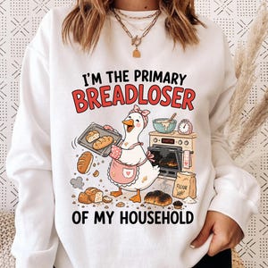 May include: White sweatshirt with a cartoon goose wearing a pink apron and holding a tray of bread. Text reads "I'M THE PRIMARY BREADLOSER OF MY HOUSEHOLD". Baking-themed design.