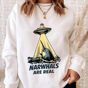 Könnte beinhalten: Weißes Sweatshirt mit einem Cartoon-Narwal, der von einem UFO entführt wird. Der Narwal ist blau, das UFO schwarz und silber. Der Text "NARWHALS ARE REAL" ist darunter gedruckt.