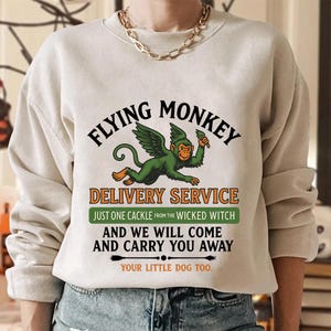 Può includere: Felpa beige con una grafica di una scimmia alata verde che tiene una bacchetta. Il testo recita "FLYING MONKEY DELIVERY SERVICE" con testo aggiuntivo sottostante. Il design include frecce e la frase "YOUR LITTLE DOG TOO."