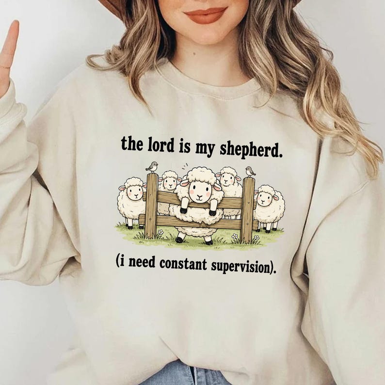 May include: Cream-colored sweatshirt featuring a cartoon sheep design. The graphic shows sheep behind a wooden fence with the text "the lord is my shepherd. (i need constant supervision)." Two small birds are perched on the fence.