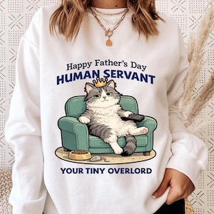 May include: White sweatshirt with a graphic of a cat wearing a crown, sitting on a teal armchair, holding a remote. Text reads "Happy Father's Day HUMAN SERVANT YOUR TINY OVERLORD". A cat food bowl and toy mouse are also shown.