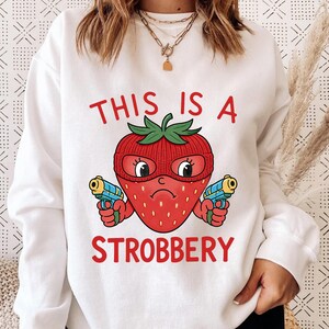 May include: White sweatshirt with the text "THIS IS A STROBBERY". The graphic features a red strawberry wearing a red knit cap and holding two blue and yellow toy guns. The strawberry has a sad expression.